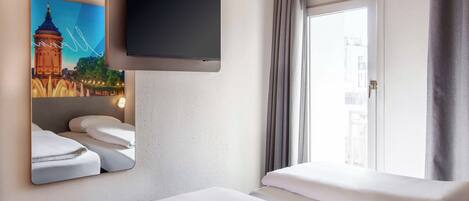 Habitación de hotel con una televisión de pantalla plana montada en la pared, una cama con ropa blanca, una ventana con cortinas y un cuadro de un puente.