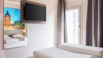 A hotel room with a bed, a flat-screen TV mounted on the wall, a window with curtains, and a wall-mounted mirror reflecting a cityscape.