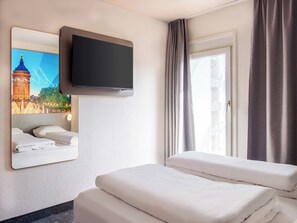 Habitación de hotel con cama, televisor de pantalla plana en la pared, ventana con cortinas y espejo adosado que refleja un paisaje urbano.