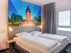 Une chambre d’hôtel comprenant un lit, une table de chevet, une lampe fixée au mur, une fenêtre avec des rideaux et un tableau représentant une tour.