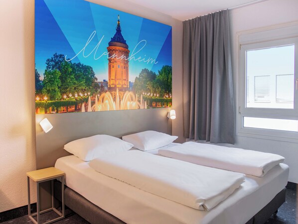 Une chambre d’hôtel comprenant un lit, une table de chevet, une lampe fixée au mur, une fenêtre avec des rideaux et un tableau représentant une tour.