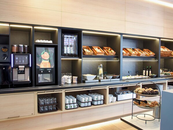 Un espace de petit-déjeuner en libre-service comprenant une machine à café, des boissons réfrigérées et une variété d’aliments.