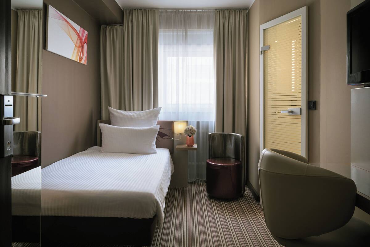 Een hotelkamer met een bed, een stoel, een televisie, een lamp en een raam met gordijnen.