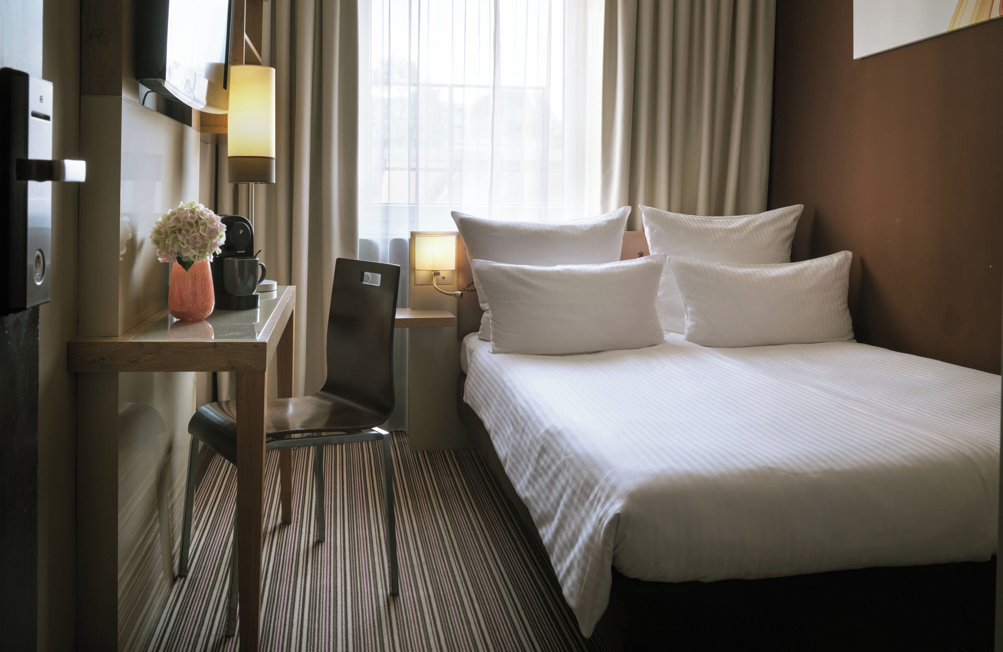Habitación de hotel con cama, escritorio, silla, lámpara, un jarrón con flores y una ventana con cortinas.