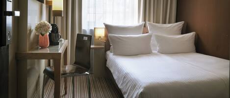Habitación de hotel con cama, escritorio, silla, lámpara, un jarrón con flores y una ventana con cortinas.