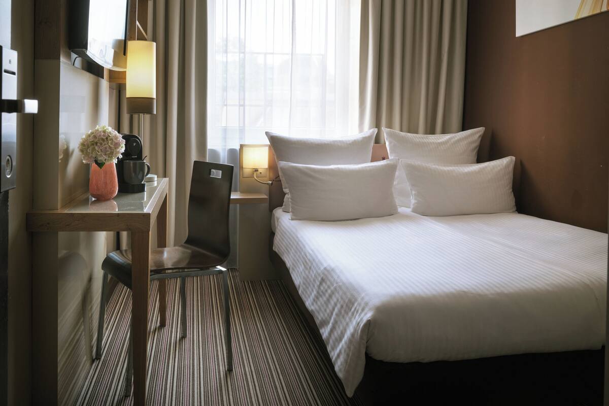 Een hotelkamer met een bed, een bureau, een stoel, een lamp, een vaas met bloemen en een raam met gordijnen.