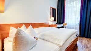 A neatly made bed with white linens and pillows, a wooden headboard, a bedside table with a lamp, and a window with curtains. - Hotel Antares (Munich)