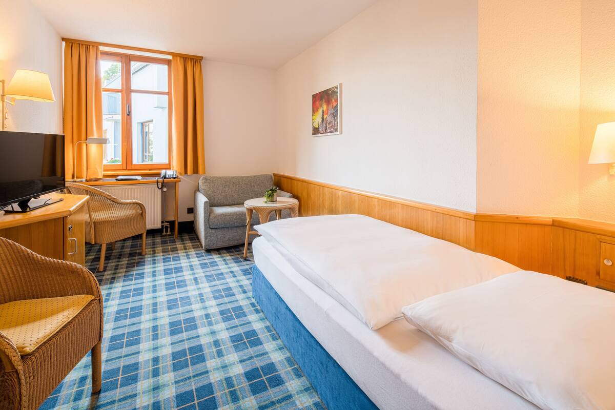 A hotel room with a checkered floor, a bed, a desk with a computer, a chair, a small table with a lamp, and a window with curtains.
