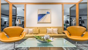 Una sala moderna con una mesa de centro de vidrio, dos sillas naranjas, un sofá con cojines estampados y un cuadro abstracto enmarcado en la pared.