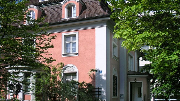 A two-story building with a red facade and a brown roof, surrounded by greenery. - Hotel Galleria (Munich)