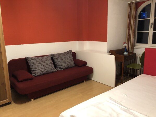 A room with a red and white wall, a red sofa with patterned cushions, a small wooden table with a lamp, and a window with curtains. - Hotel Galleria (Munich)