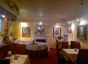 A dining area with round tables and chairs, a fireplace, and various wall decorations. - Hotel Galleria (Munich)