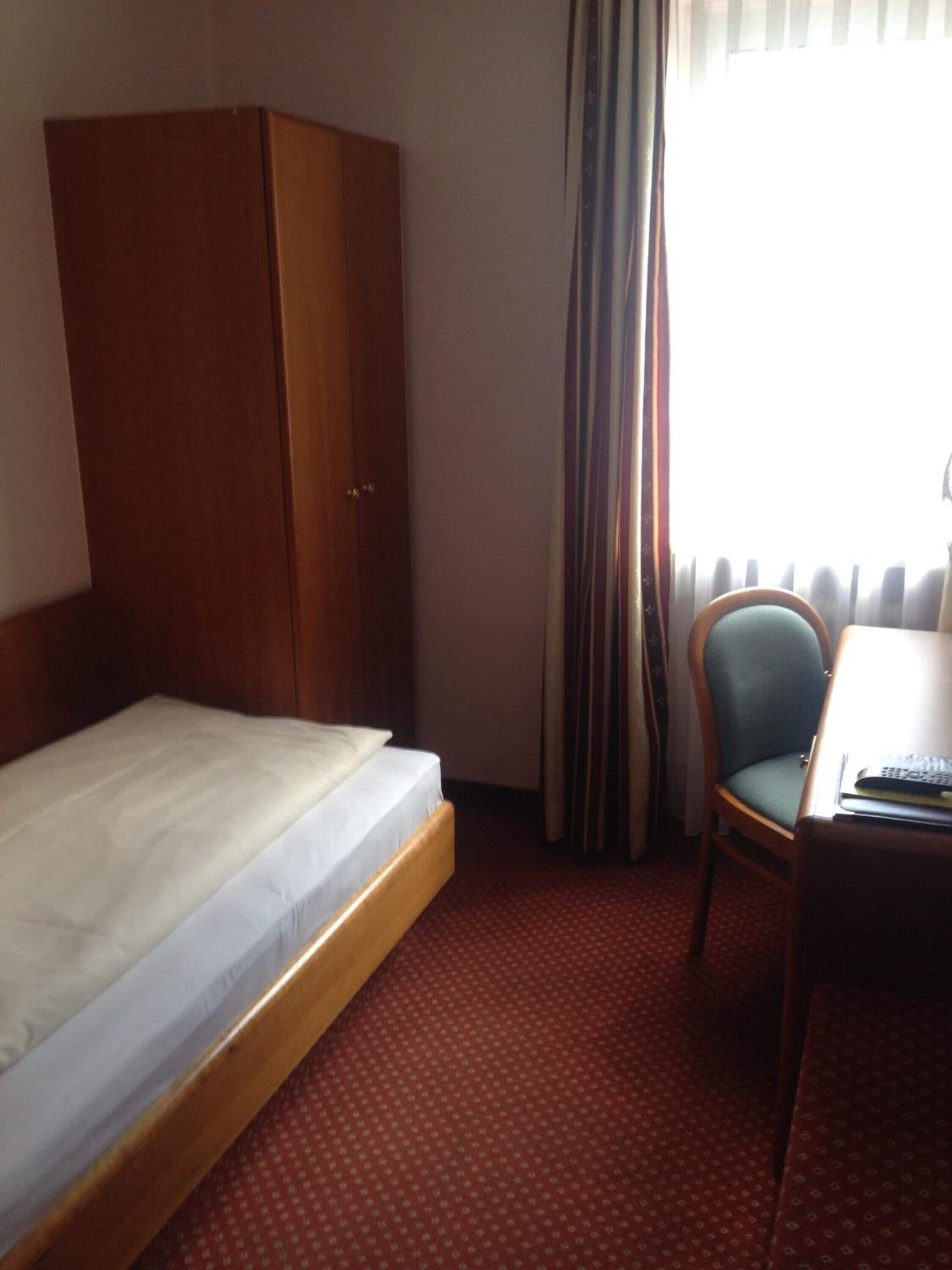 Une chambre d’hôtel avec un lit simple, une armoire en bois, un bureau avec une chaise et une fenêtre avec des rideaux.