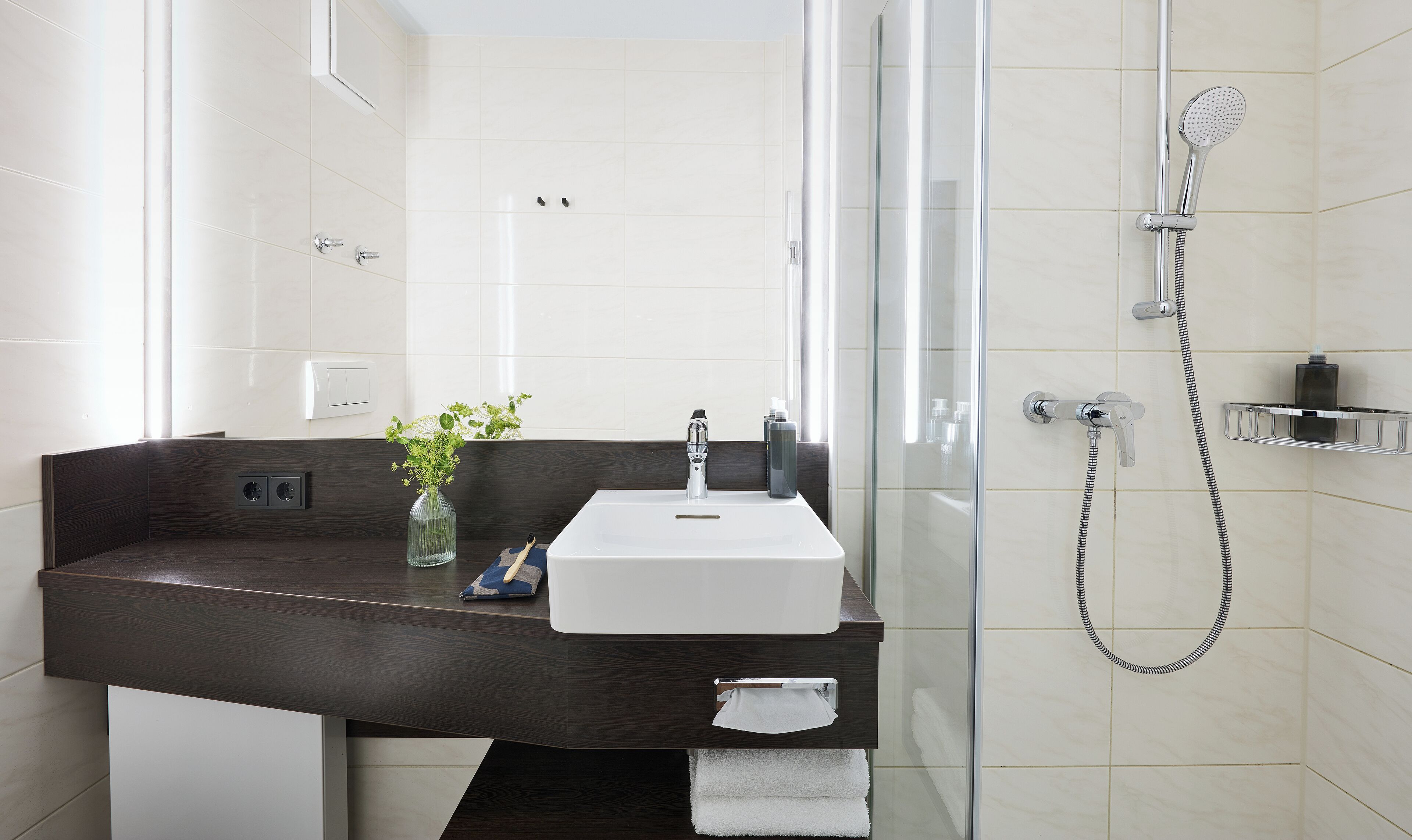 Un baño moderno con tocador de madera oscura, un lavabo blanco, cabina de ducha de vidrio y un perchero para toallas.