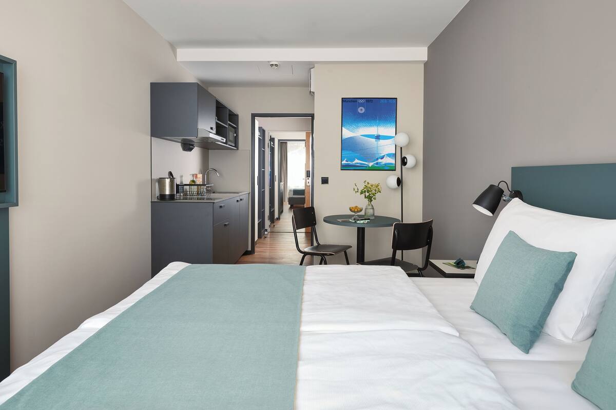 A hotel room with a bed, a desk, a chair, a small table, a TV, and a painting on the wall.