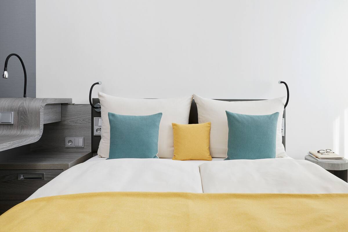 A neatly made bed with a yellow blanket, two pillows, and a bedside table with a book and glasses.
