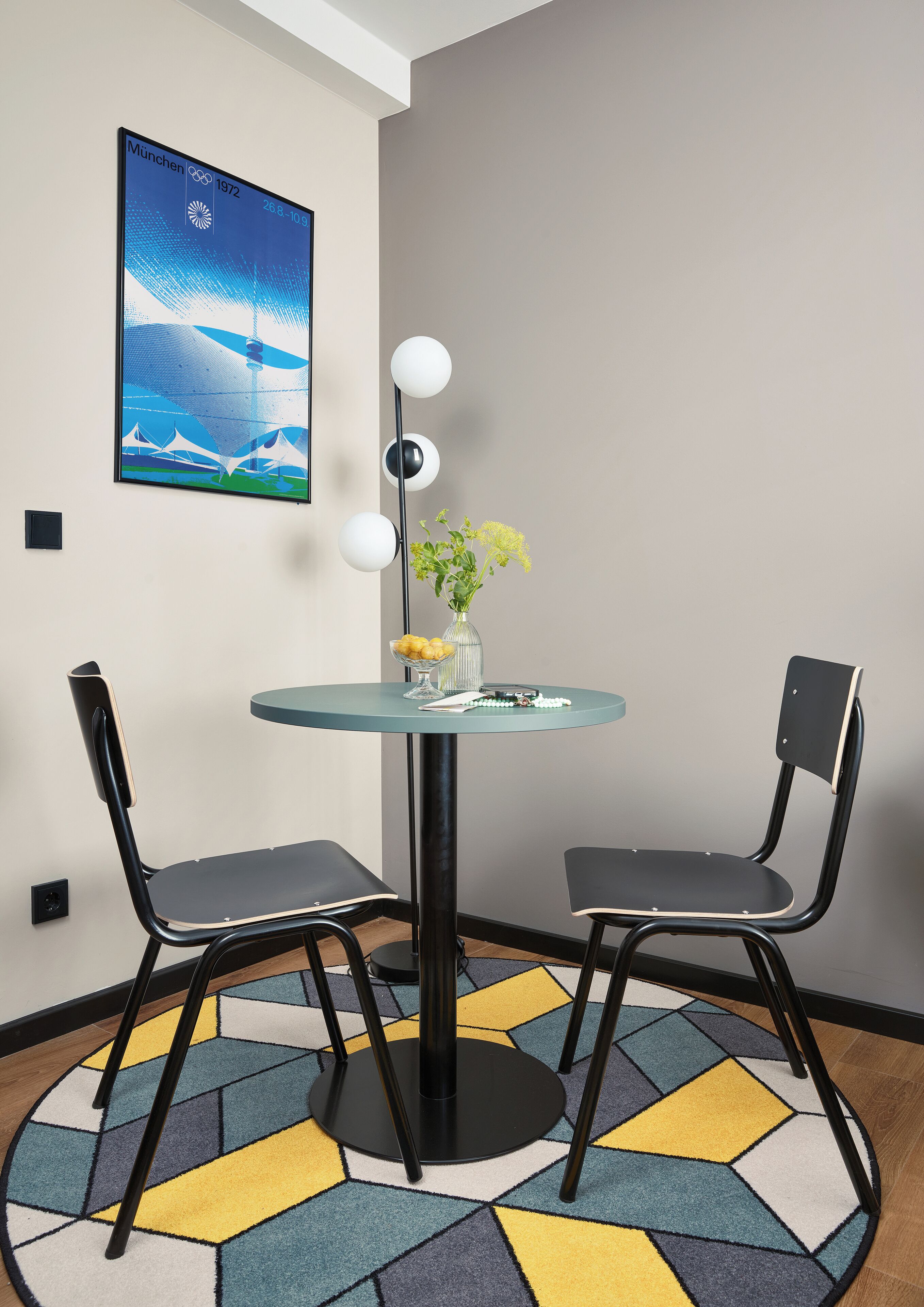 A modern dining area with a glass table, two black chairs, a colorful rug, a vase with flowers, and a framed picture on the wall.
