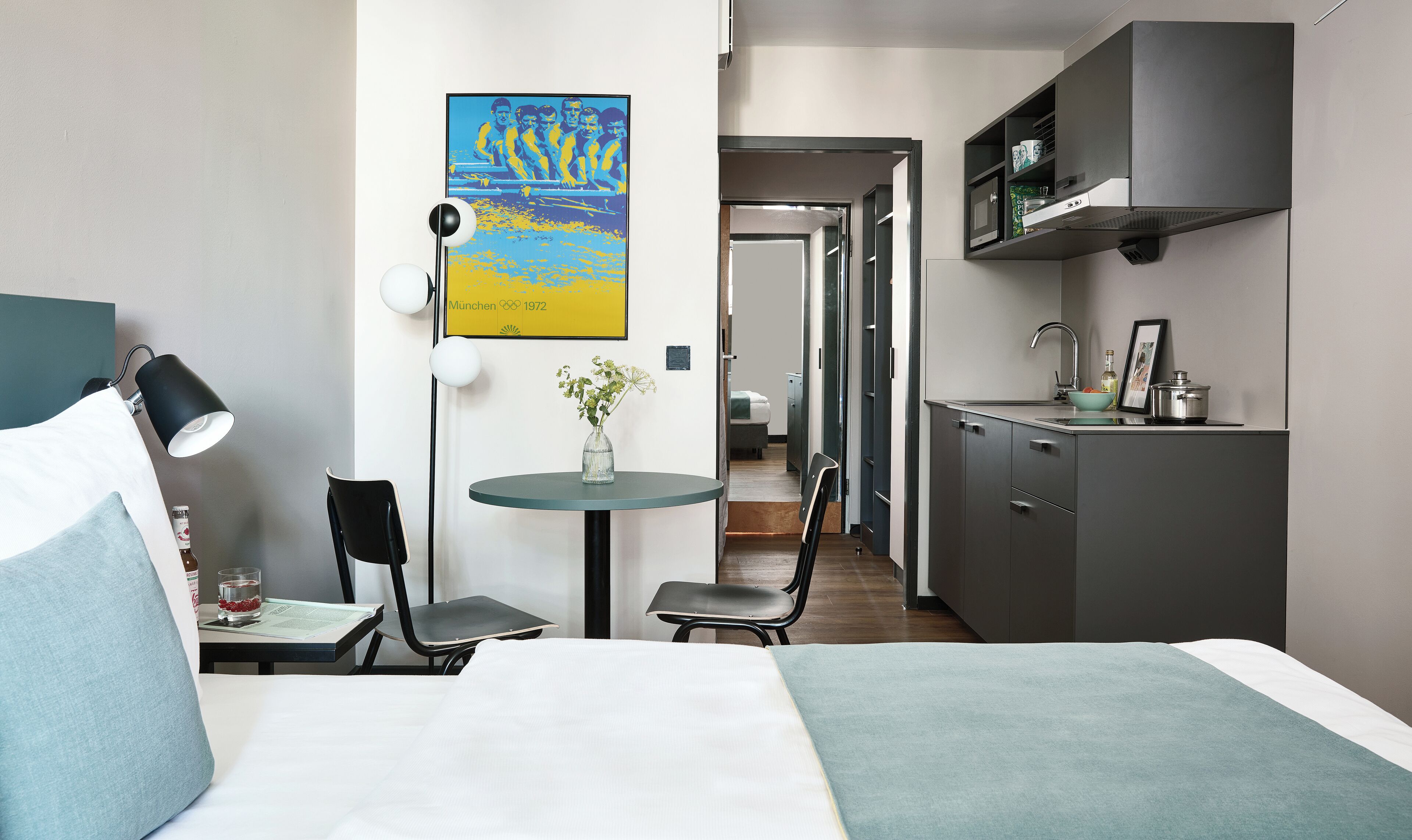 A compact hotel room with a bed, a small table with a chair, a desk with a chair, a kitchenette, and a framed picture on the wall.