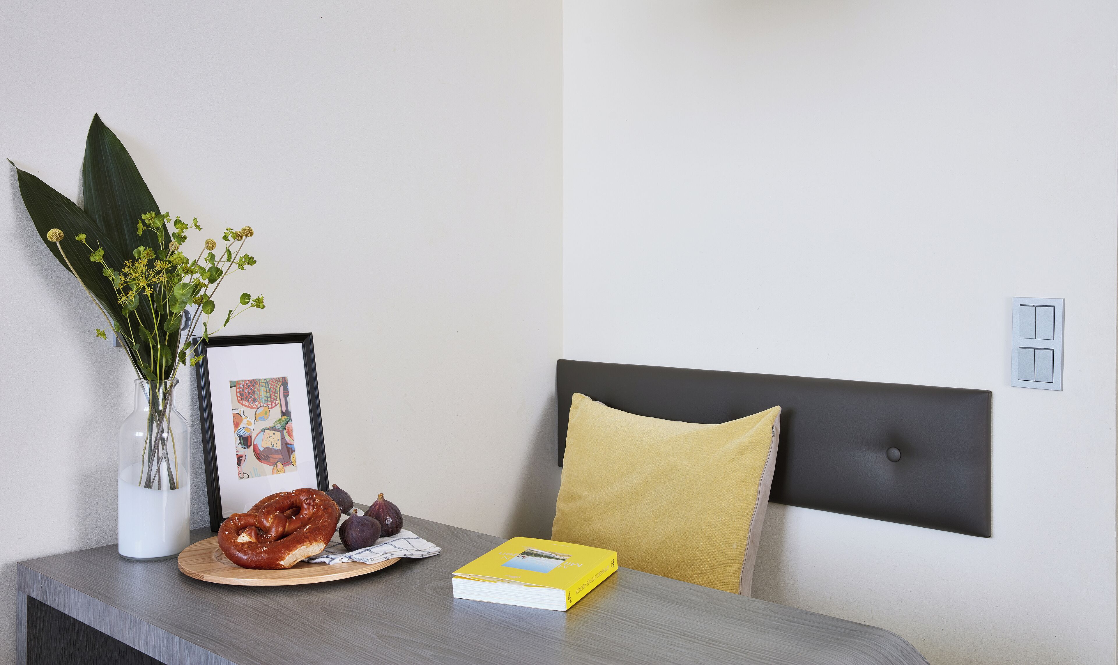 A room with a bed, a bedside table, a vase with flowers, a framed picture, a book, and a plate with food.