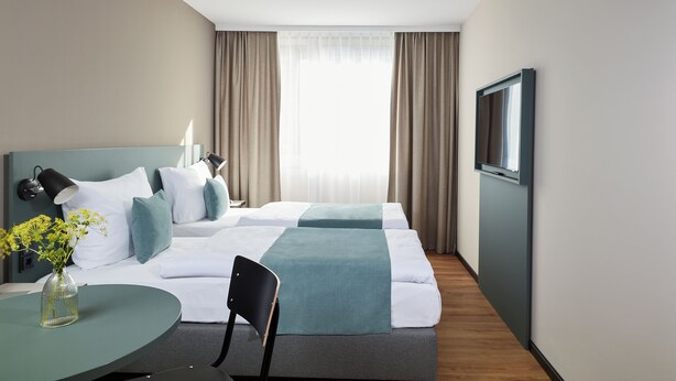 Una habitación de hotel con cama, escritorio, silla, televisor y ventana con cortinas.