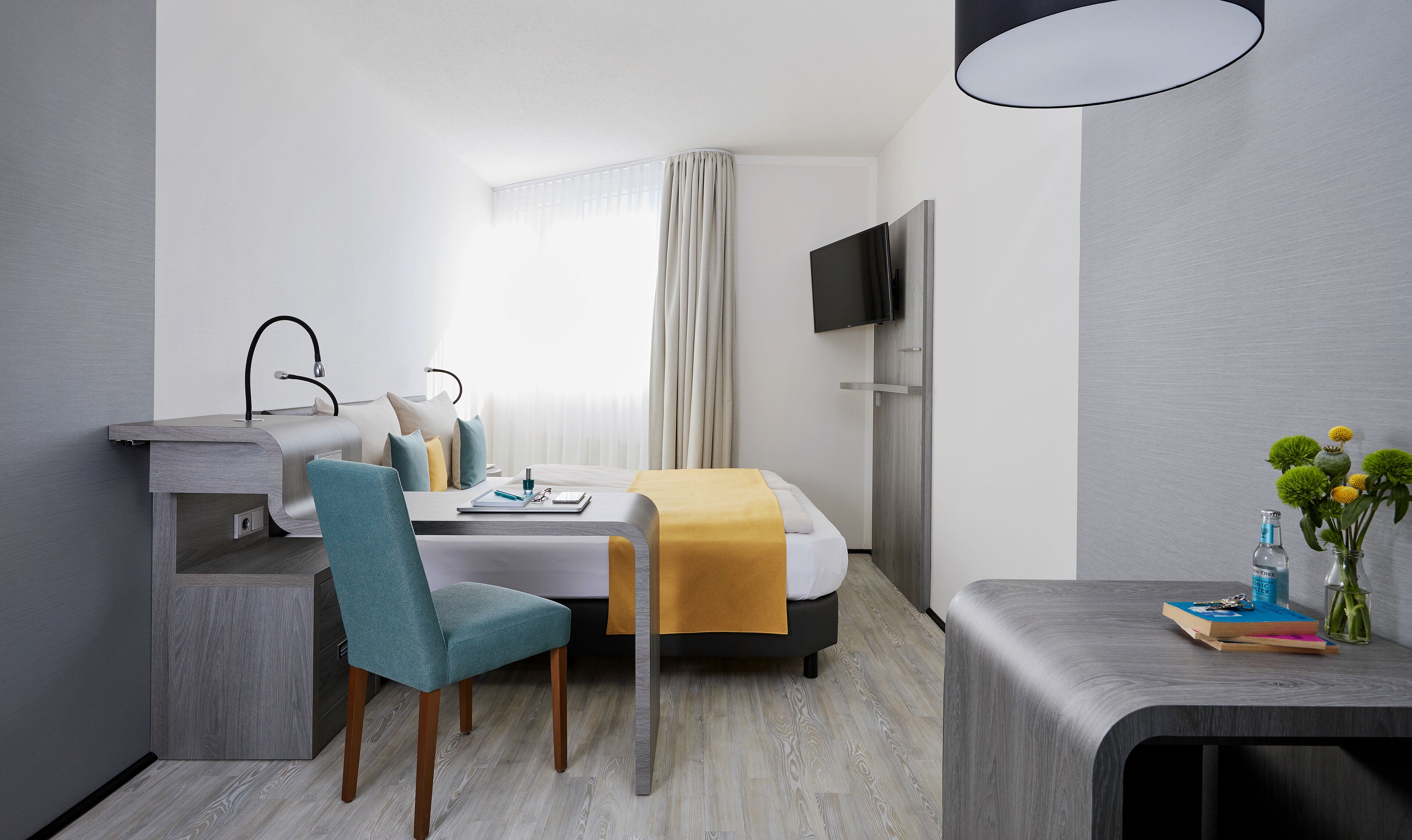 Ein modernes Hotelzimmer mit Bett, Schreibtisch, Sessel, Fernseher und Nachttisch mit Blumenvase.