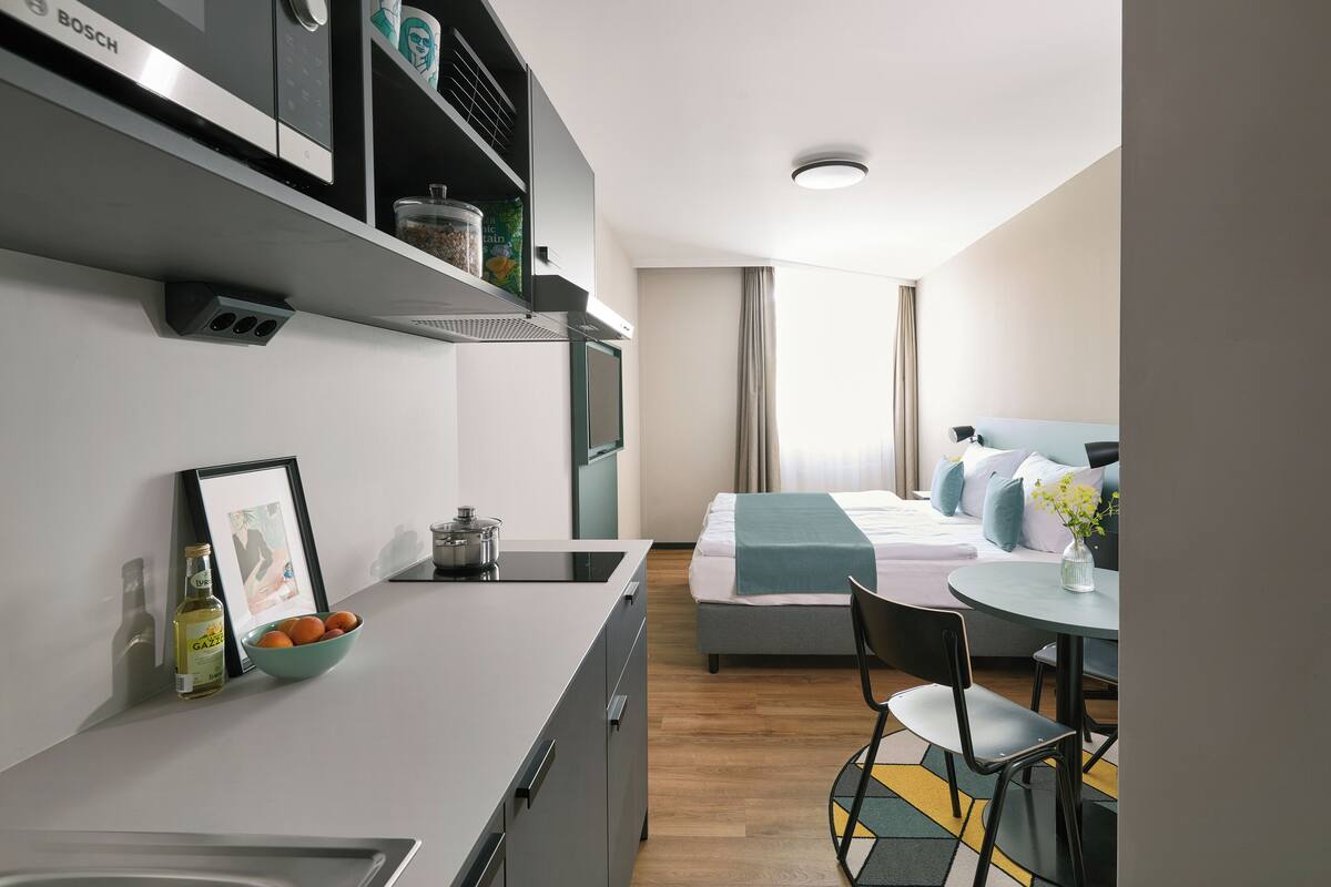 A modern hotel room with a kitchenette, a bed with a blue blanket, a dining area with a table and chairs, and a window with curtains.