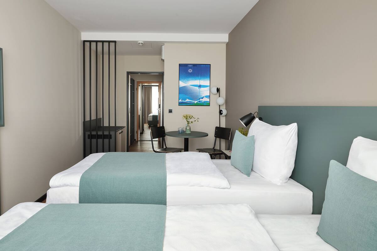 A hotel room with two beds, a dining table, and a view of the hallway.