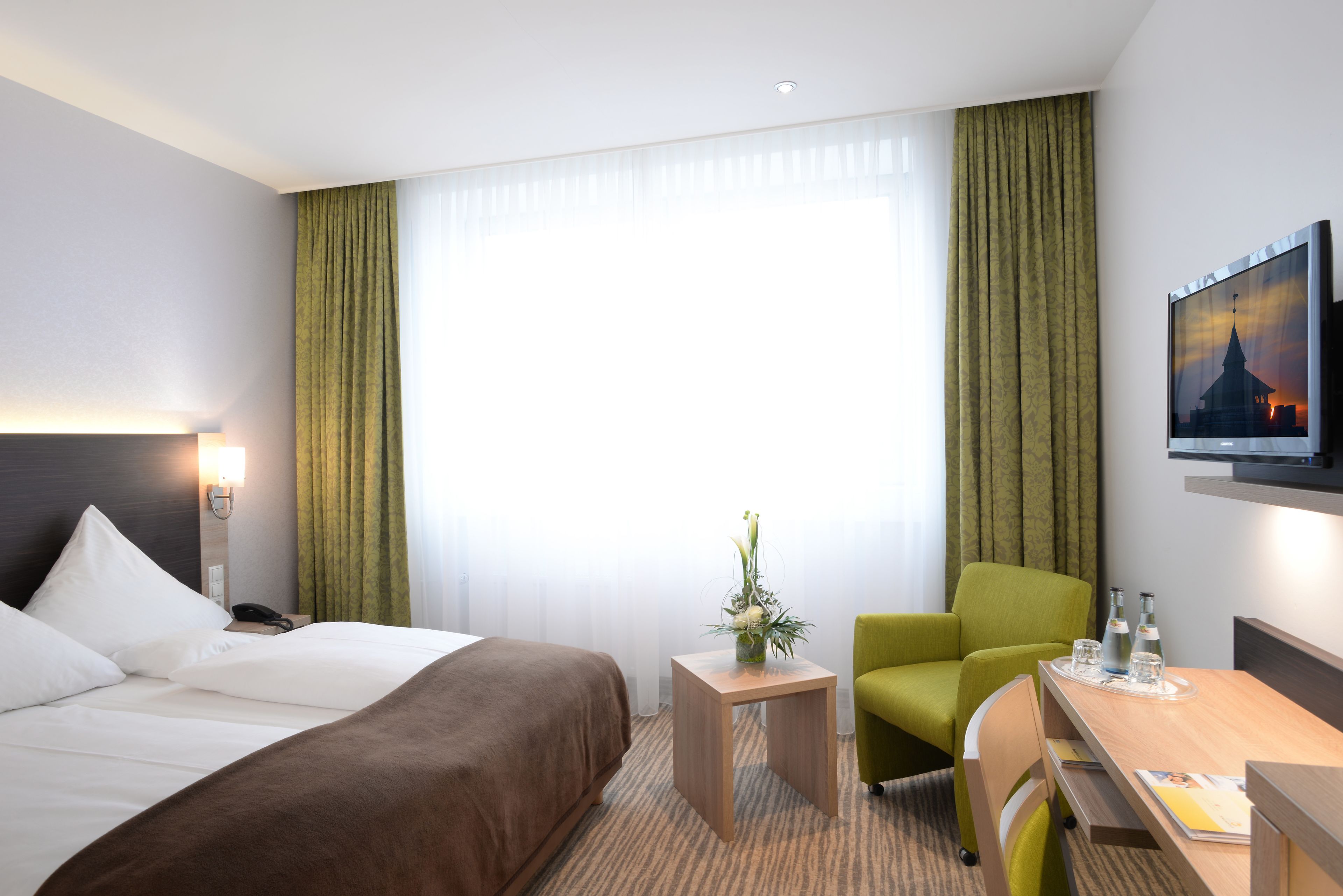 Ein Hotelzimmer mit Bett, Schreibtisch, Stuhl, einem kleinen Tisch mit Vase und einem Bild an der Wand.