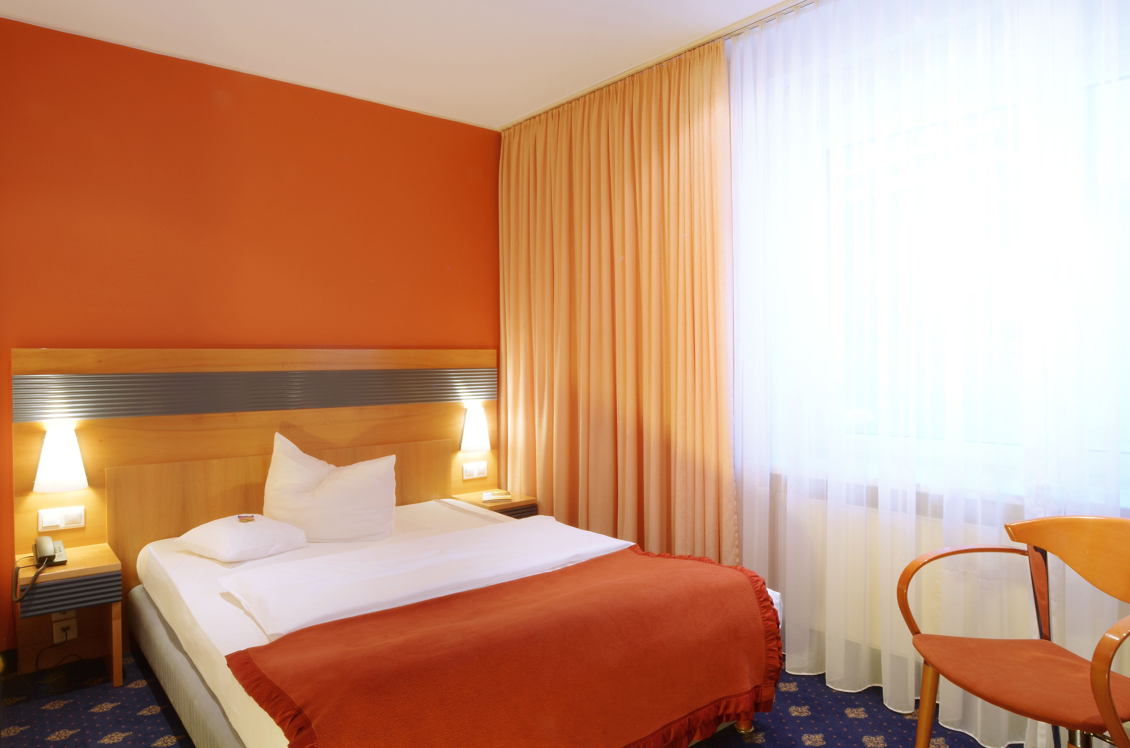 A hotel room with a large bed, two bedside lamps, a wooden headboard, a red throw blanket, a wooden chair, and a window with sheer curtains.