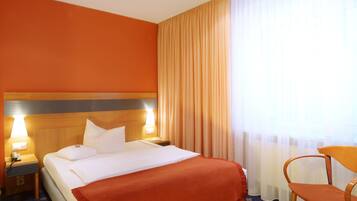 A hotel room with a large bed, two bedside lamps, a wooden headboard, a red throw blanket, a wooden chair, and a window with sheer curtains.