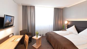A hotel room with a bed, a desk, a chair, a small table with a vase, and a television on the wall.