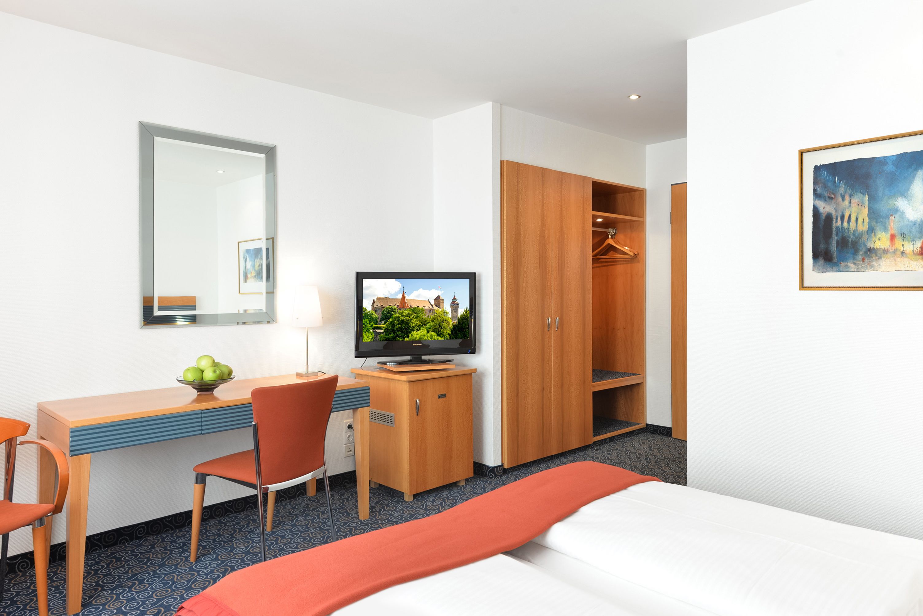 A hotel room with a bed, a desk with a chair, a television, a wardrobe, and a painting on the wall.