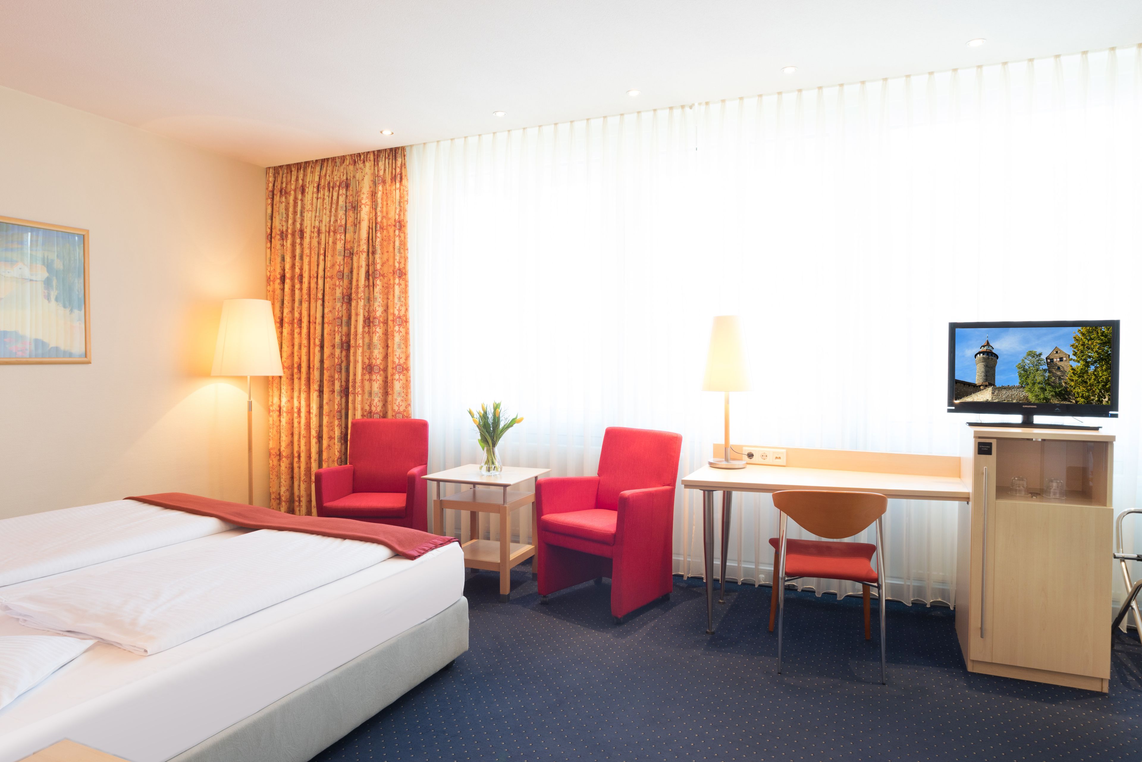 A hotel room with a bed, two red chairs, a desk, a television, and a small table with a lamp.