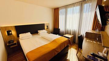 Ein Hotelzimmer mit einem großen Bett, Nachttischen, einem Schreibtisch, einem Sessel und Vorhängen.