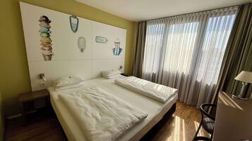 A hotel room with two beds, a large window with curtains, a bedside table, and artwork on the wall.