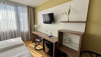 A hotel room with a bed, a desk with a chair, a television mounted on the wall, and a small shelf with a vase.