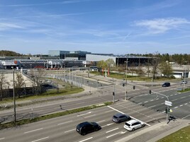 Una carretera con varios carriles, vehículos, un paso de peatones y un gran edificio industrial al fondo.