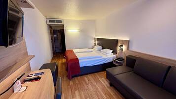 A compact hotel room with a bed, a desk, a chair, a sofa, and a TV.