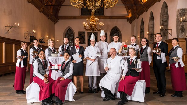 A group of waitstaff in a formal dining setting with a chandelier and arched windows.