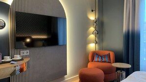 Une chambre d’hôtel moderne avec un grand téléviseur à écran plat fixé au mur, une table de chevet en bois avec une tasse et une soucoupe, un fauteuil orange avec un coussin à rayures, une petite table ronde et un lit avec une couette blanche.