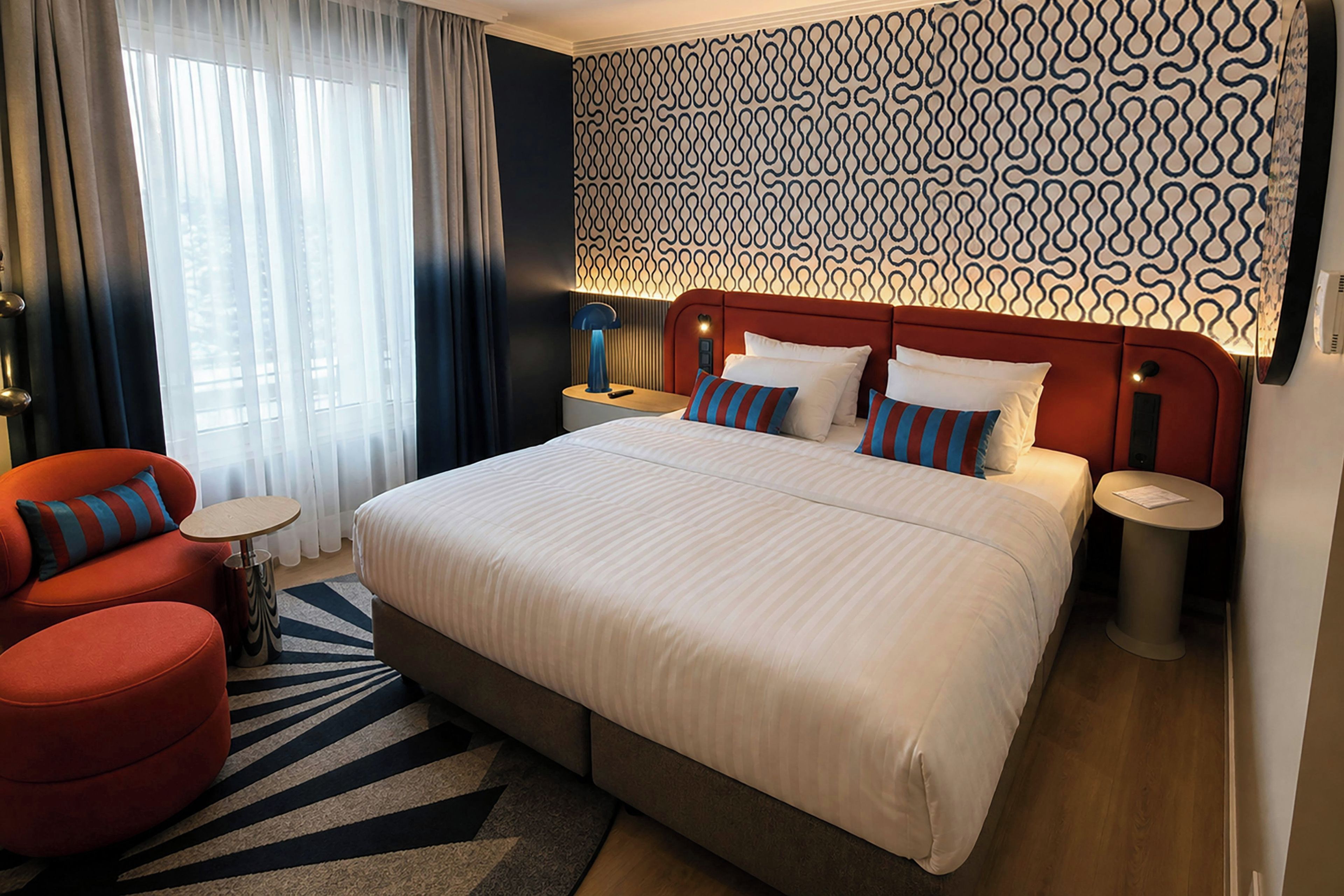 A hotel room with a large bed, a red armchair, a small round table, and a patterned wall.
