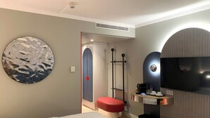 Une chambre d’hôtel moderne avec un lit, une œuvre d’art murale ronde, un banc rouge, une télévision et une petite étagère avec quelques objets.