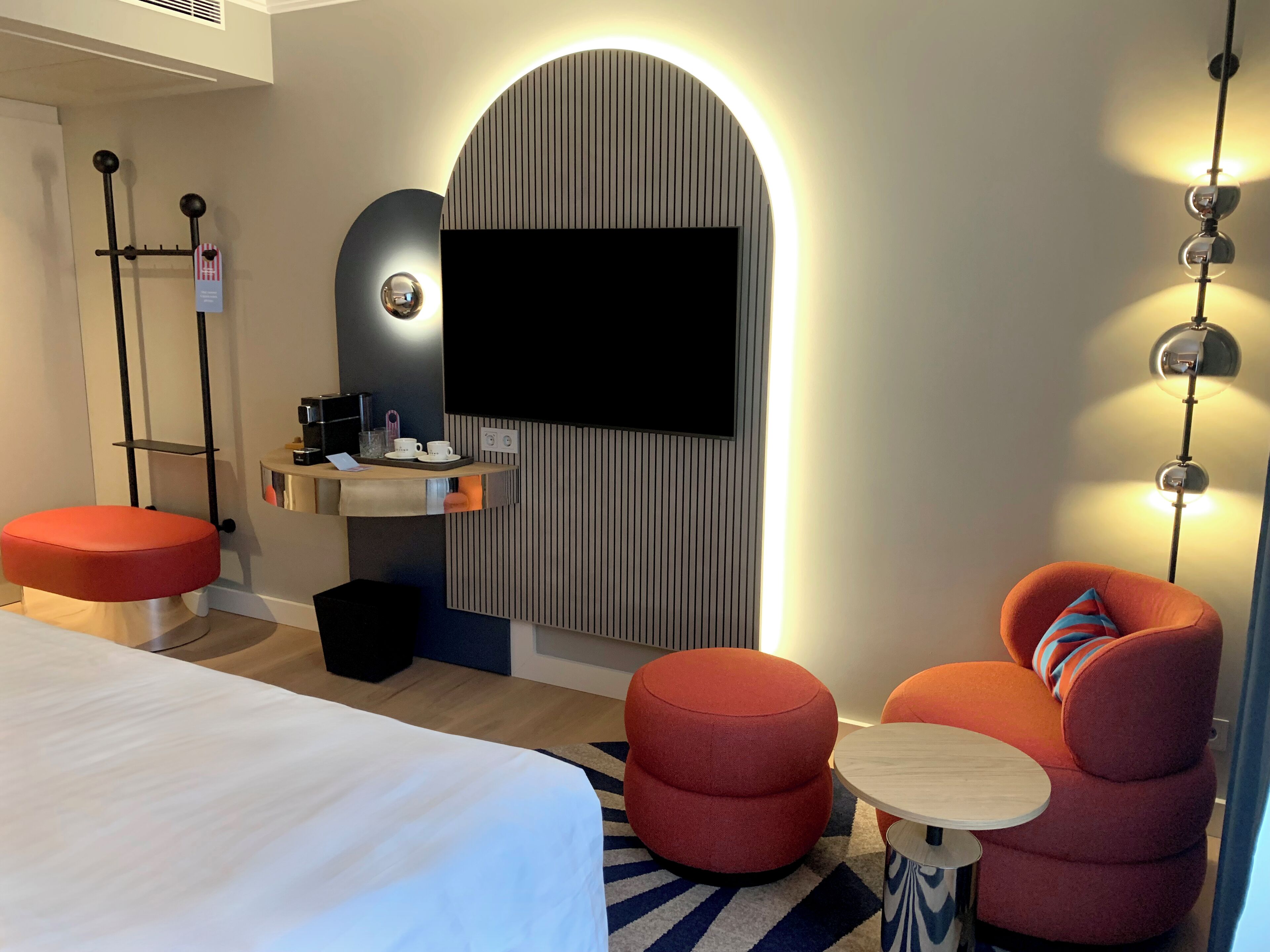 A modern hotel room with a bed, a seating area with a round table, a coffee station, and a television.