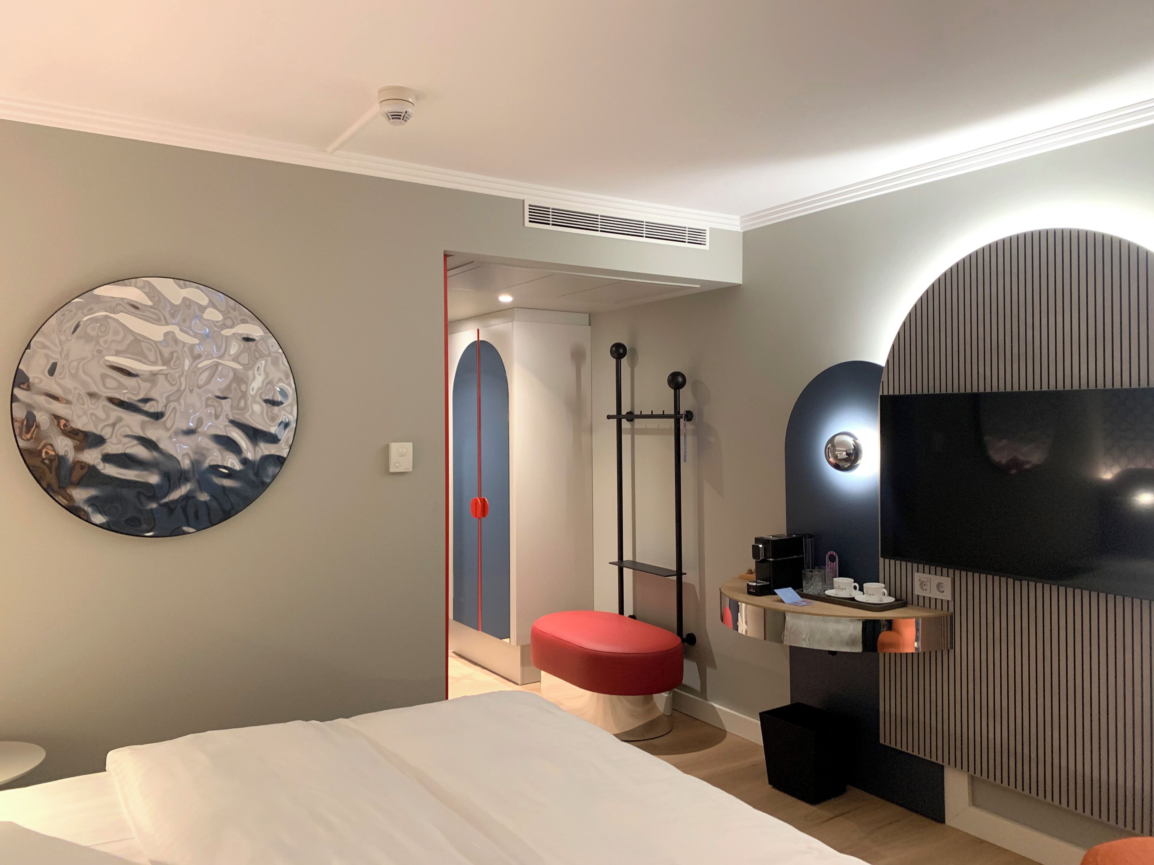 A modern hotel room with a bed, a round wall art piece, a red bench, a TV, and a small table with a cup and a remote control.