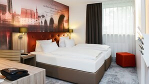 A hotel room with a large bed, a desk, a telephone, and a red ottoman. - Likestay Regensburg (Regensburg)