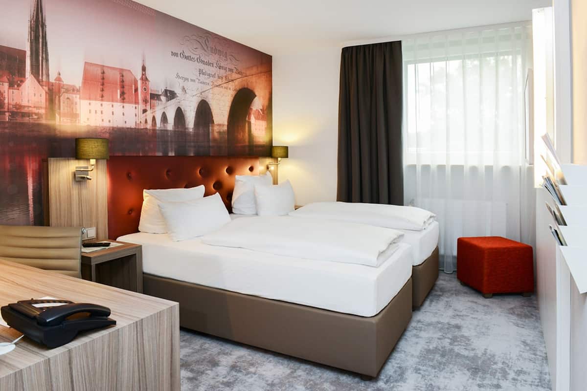 Une chambre d’hôtel avec un grand lit, un bureau, un téléphone et un pouf rouge.