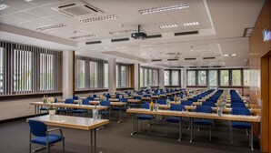 A conference room with rows of tables and chairs, large windows, and a projector on the ceiling. - Likestay Regensburg (Regensburg)