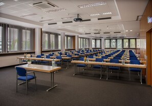 Un salón de conferencias con filas de mesas y sillas, ventanas amplias y un proyector en el techo.