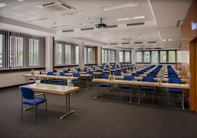 Un salón de conferencias con filas de mesas y sillas, ventanas amplias y un proyector en el techo.