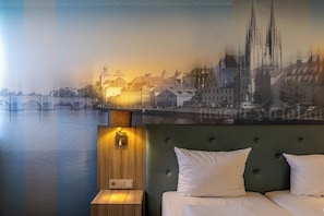 Une chambre d’hôtel avec un lit, une table de chevet, une lampe fixée au mur et une fresque représentant un paysage urbain.